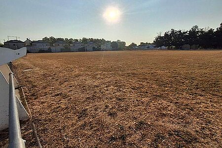 Terrain de football sous un ciel bleu azur avec un grand soleil, avec une pelouse jaunie et clairsem&eacute;e.