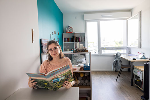 Etudiante assise &agrave; la table de son logement &eacute;tudiant. Un burerau et des &eacute;tag&egrave;res sont visibles &agrave; l'arri&egrave;re-plan.